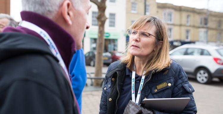 Woman with a Healthwatch lanyard and clipboard speaking to a member of the public outside