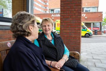 Two older women sitting on a bench talking outside A&E