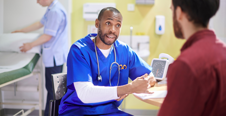 A medical professional wearing blue scrubs, talking to a patient at a table in a hospital. Behind them, a second medical professional is making up a hospital bed.