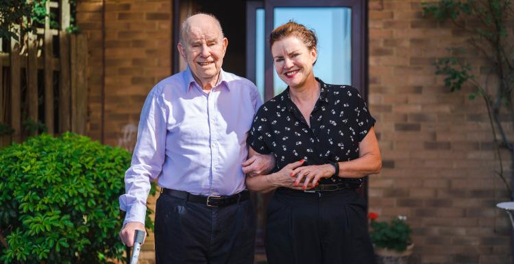 A father and daughter walking outside a house, arm in arm. The father is using a walking stick.