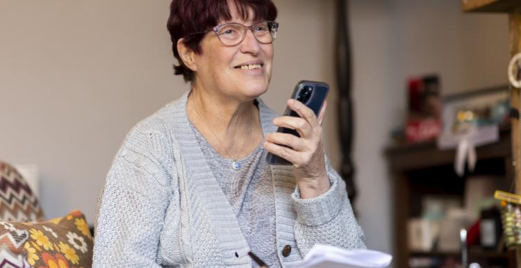 A smiling woman holding a smartphone and some pieces of paper.