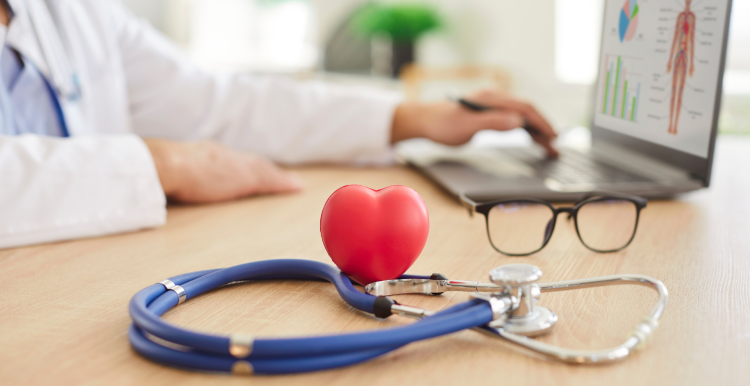 Stethoscope with a red heart-shaped object and glasses on a desk in a doctor's office. A doctor is working on a laptop in the background.