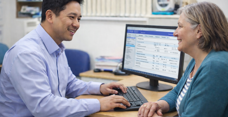 A male doctor talking to a female patient in a doctor's office. The doctor is typing on a computer.