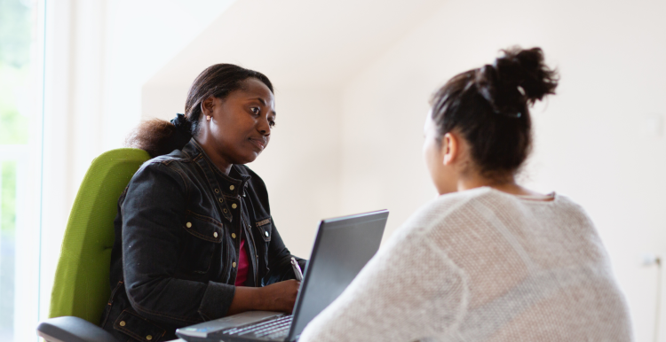 A woman sitting behind a laptop, looking thoughtful. She is talking to another woman who is sitting across from her.