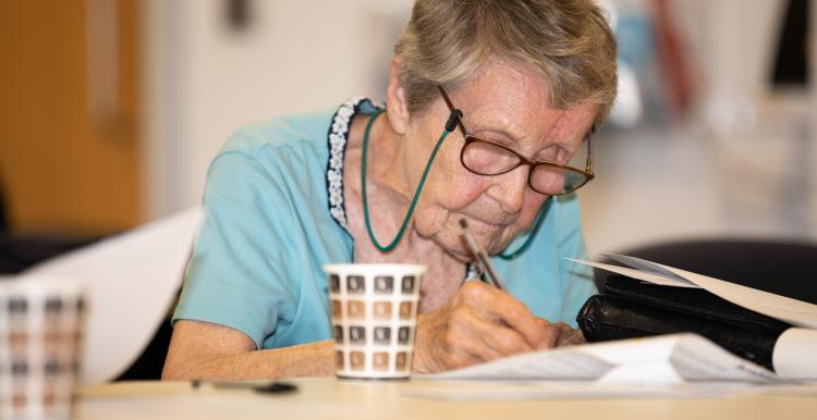 An elderley woman at a table writing on a piece of paper