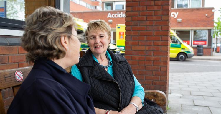 Two older women sitting on a bench talking outside A&E