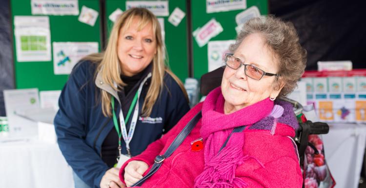 Elderly woman in a wheelchair smiling at the camera at an outdoor event in front of a Healthwatch stand