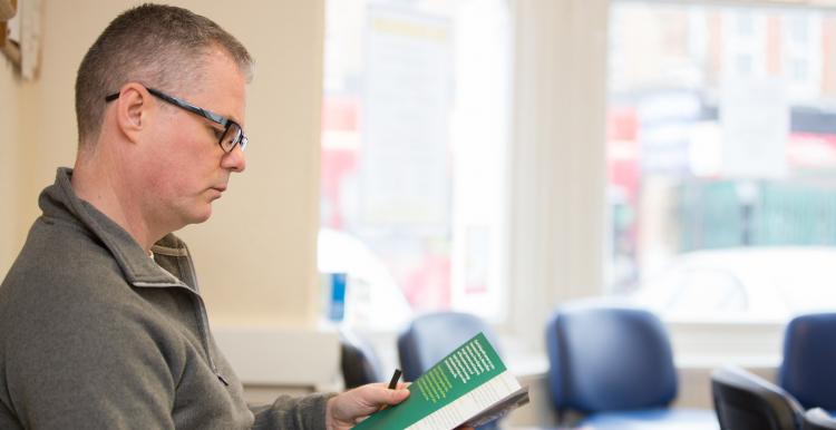 Middle aged man sitting in empty reception area of a GP reading a leaflet