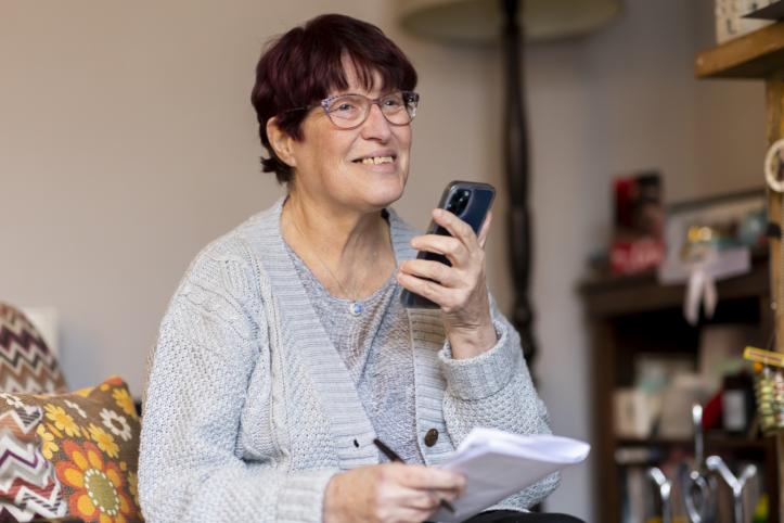 A smiling woman holding a smartphone and some pieces of paper.