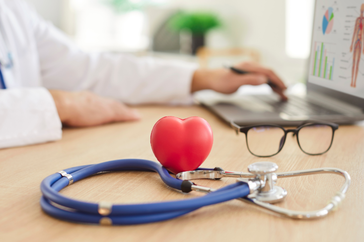 Stethoscope with a red heart-shaped object and glasses on a desk in a doctor's office. A doctor is working on a laptop in the background.
