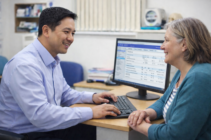 A male doctor talking to a female patient in a doctor's office. The doctor is typing on a computer.
