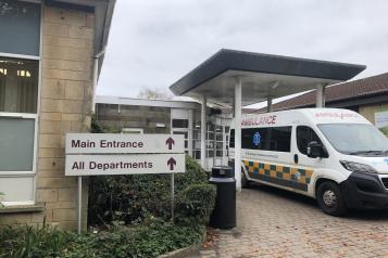 'Main Entrance' and 'All Departments' signs at the entrance to John Stacy ward. An ambulance is parked in front of the doors.