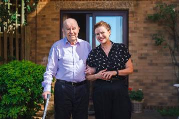 A father and daughter walking outside a house, arm in arm. The father is using a walking stick.