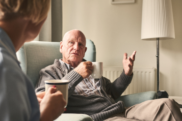 An older men sitting in an arm chair, drinking a cup of tea and talking to a carer.