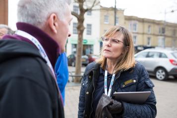 Woman with a Healthwatch lanyard and clipboard speaking to a member of the public outside