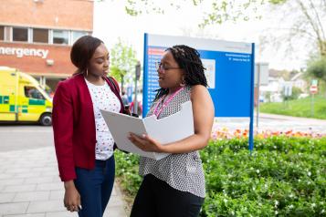 Two women outside a hospital - one has a healthwatch lanyard and is holding a clipboard speaking to the other woman as though asking questions