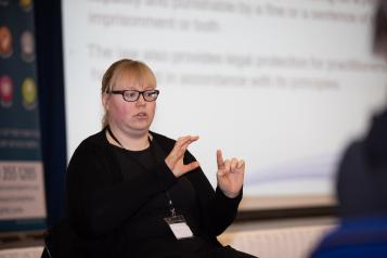Woman sitting down at an event doing British Sign Language for deaf and hard of hearing delegates