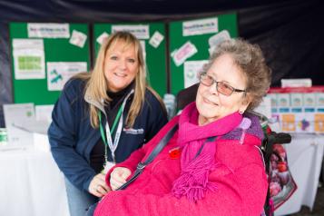 Elderly woman in a wheelchair smiling at the camera at an outdoor event in front of a Healthwatch stand