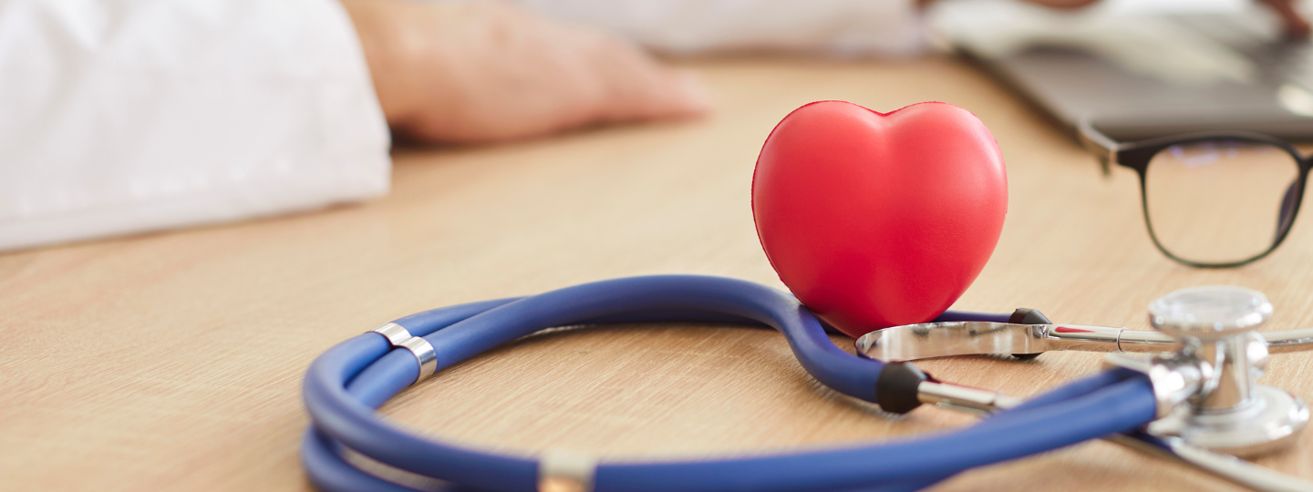 Stethoscope with a red heart-shaped object and glasses on a desk in a doctor's office. A doctor is working on a laptop in the background.