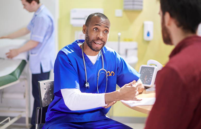 A medical professional wearing blue scrubs, talking to a patient at a table in a hospital. Behind them, a second medical professional is making up a hospital bed.