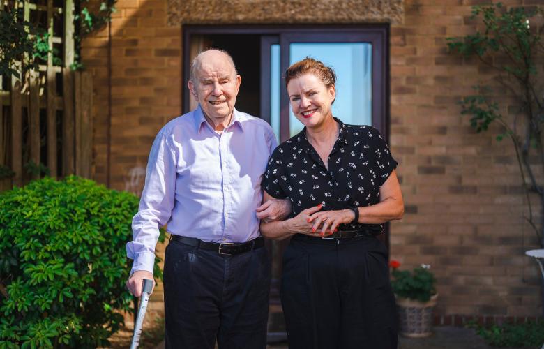 A father and daughter walking outside a house, arm in arm. The father is using a walking stick.