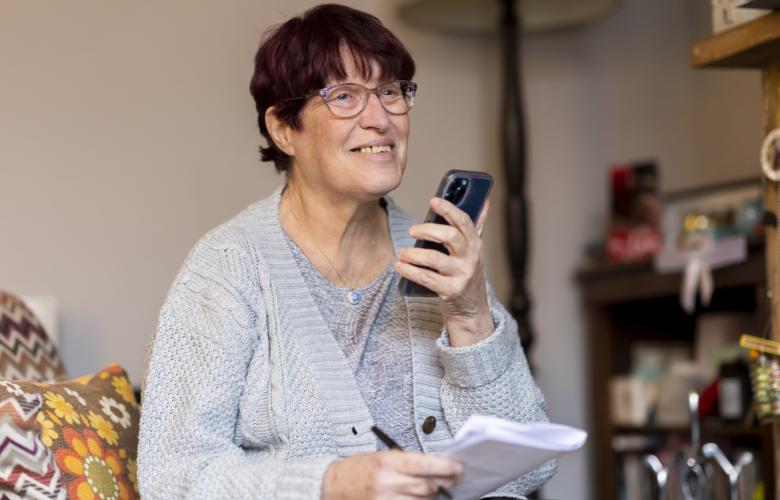A smiling woman holding a smartphone and some pieces of paper.