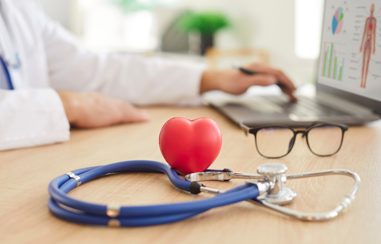Stethoscope with a red heart-shaped object and glasses on a desk in a doctor's office. A doctor is working on a laptop in the background.