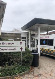 'Main Entrance' and 'All Departments' signs at the entrance to John Stacy ward. An ambulance is parked in front of the doors.