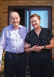 A father and daughter walking outside a house, arm in arm. The father is using a walking stick.