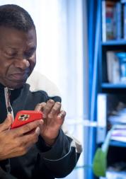 A man sitting at a desk in front of a bookcase, looking down at a mobile phone.