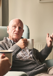 An older men sitting in an arm chair, drinking a cup of tea and talking to a carer.